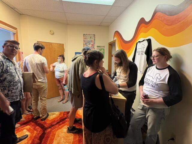 People stand in a hallway at a social event, some wearing matching white shirts with a logo, next to a colorful wavy mural and an orange-red rug.