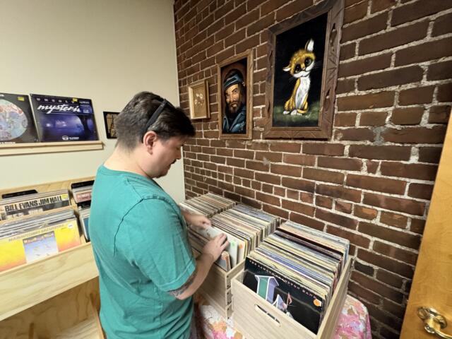 Man in teal shirt browses vinyl records in a brick-walled shop with framed art on the wall behind him
