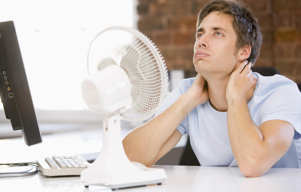 Young man sits at a desk with a white desk fan and monitor, touching his neck in apparent discomfort.