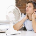 Young man sits at a desk with a white desk fan and monitor, touching his neck in apparent discomfort.