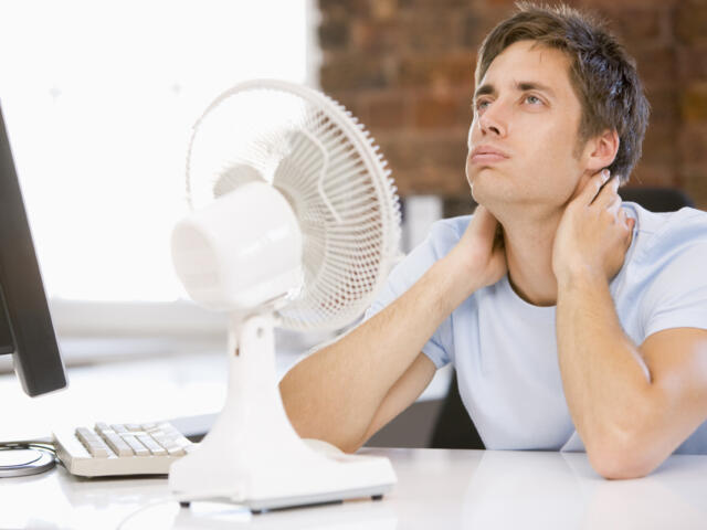 Young man sits at a desk with a white desk fan and monitor, touching his neck in apparent discomfort.