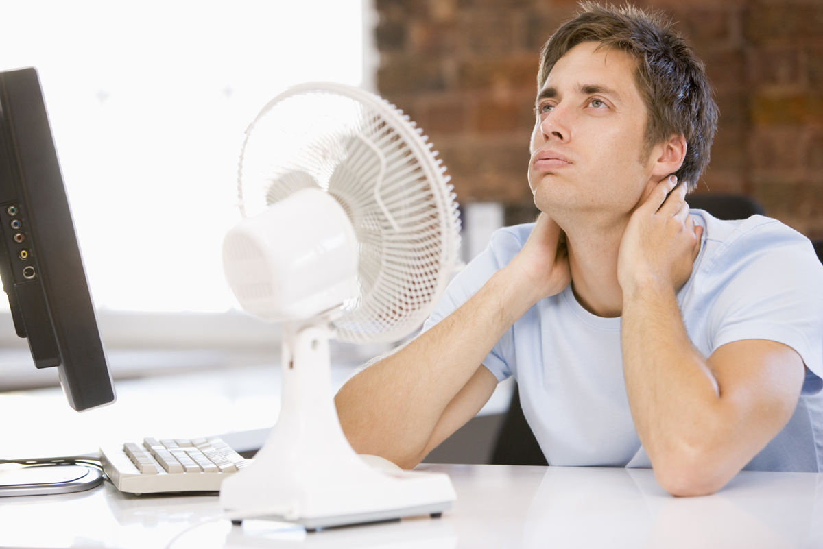 Young man sits at a desk with a white desk fan and monitor, touching his neck in apparent discomfort.