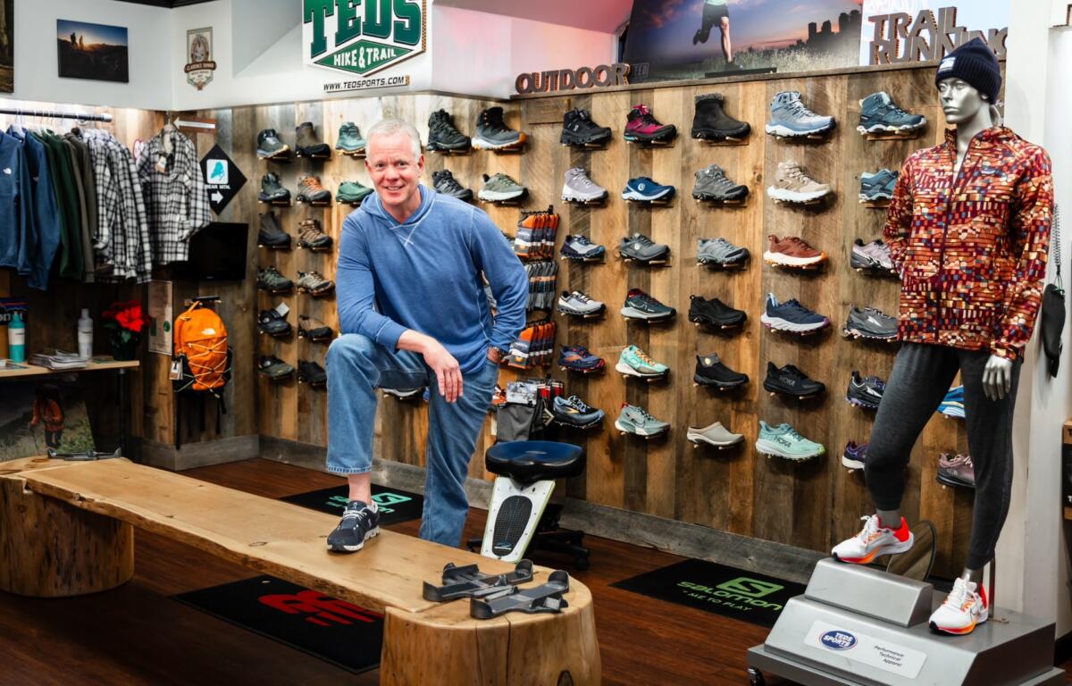 Man in a blue hoodie posing in a hiking shoe store with wall displays of athletic shoes behind him.