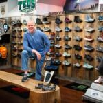 Man in a blue hoodie posing in a hiking shoe store with wall displays of athletic shoes behind him.