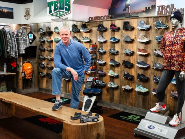 Man in a blue hoodie posing in a hiking shoe store with wall displays of athletic shoes behind him.