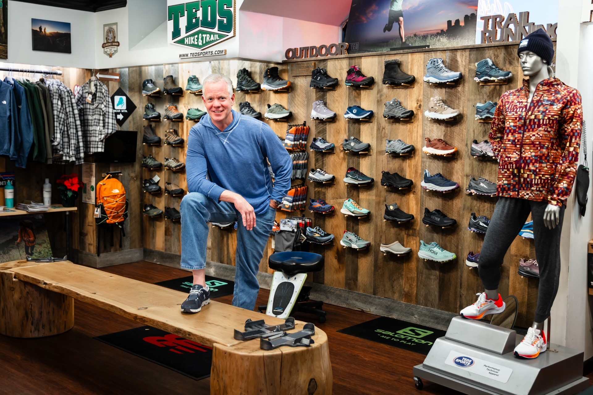 Man in a blue hoodie posing in a hiking shoe store with wall displays of athletic shoes behind him.